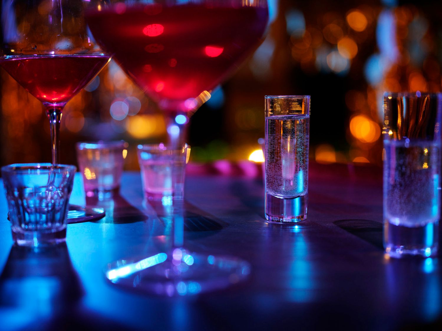 Colorful cocktail glasses and shot glasses glow under blue and pink bar lighting at night.