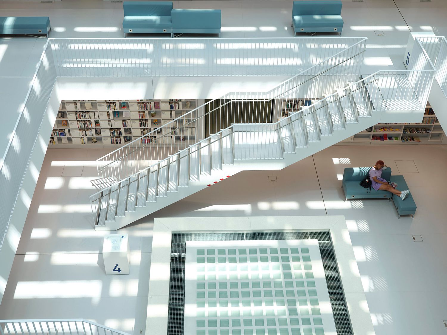 Modern white library interior with grand staircase, bookshelves, and natural light casting geometric shadows on the floor.