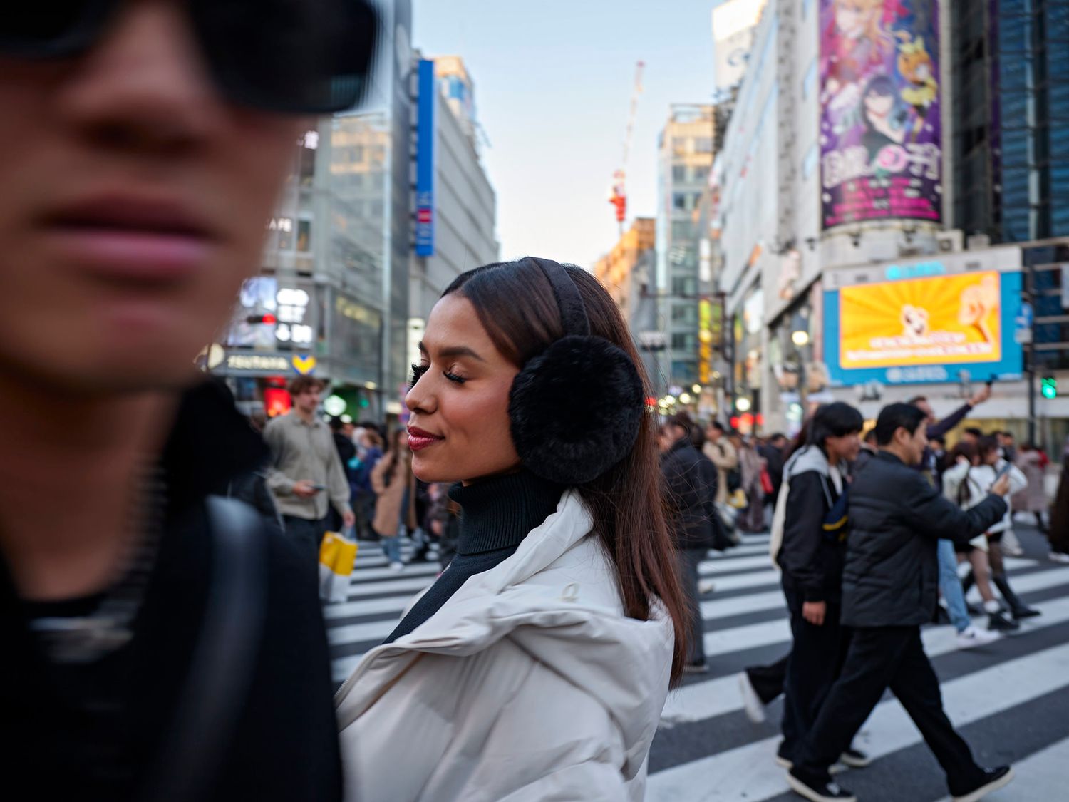 Woman wearing earmuffs crosses busy urban intersection in Tokyo, surrounded by crowds and illuminated city billboards.
