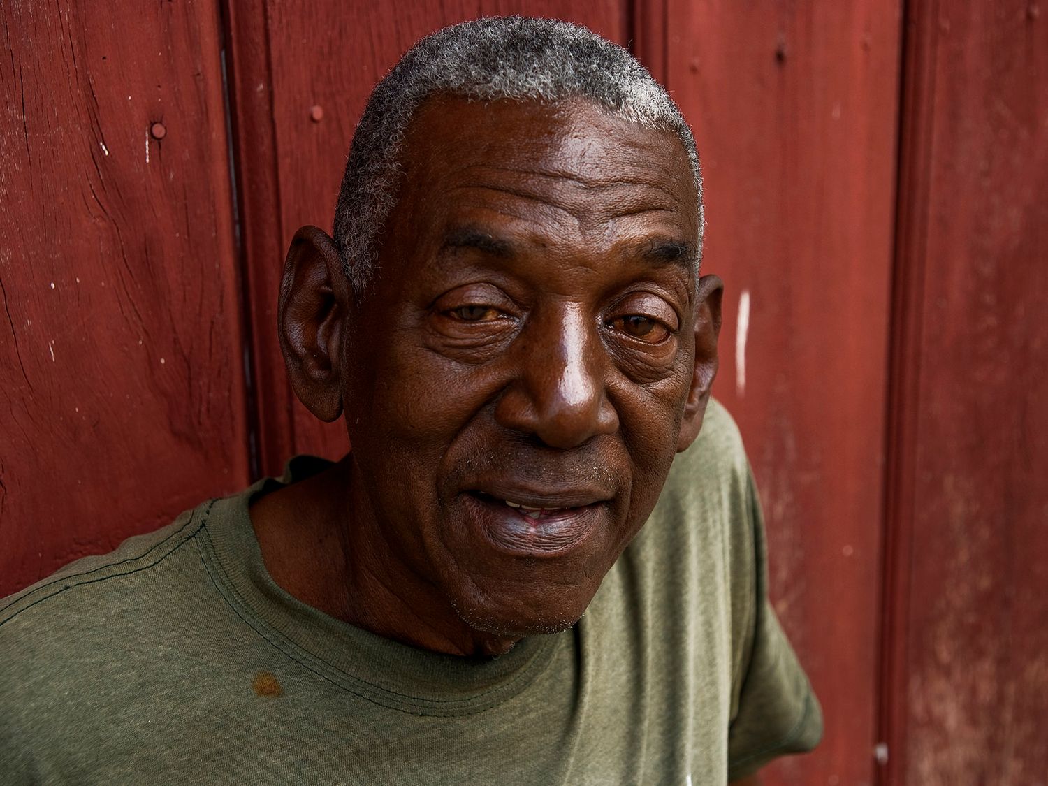 Elderly Black man in green t-shirt smiling warmly against a red wooden background.