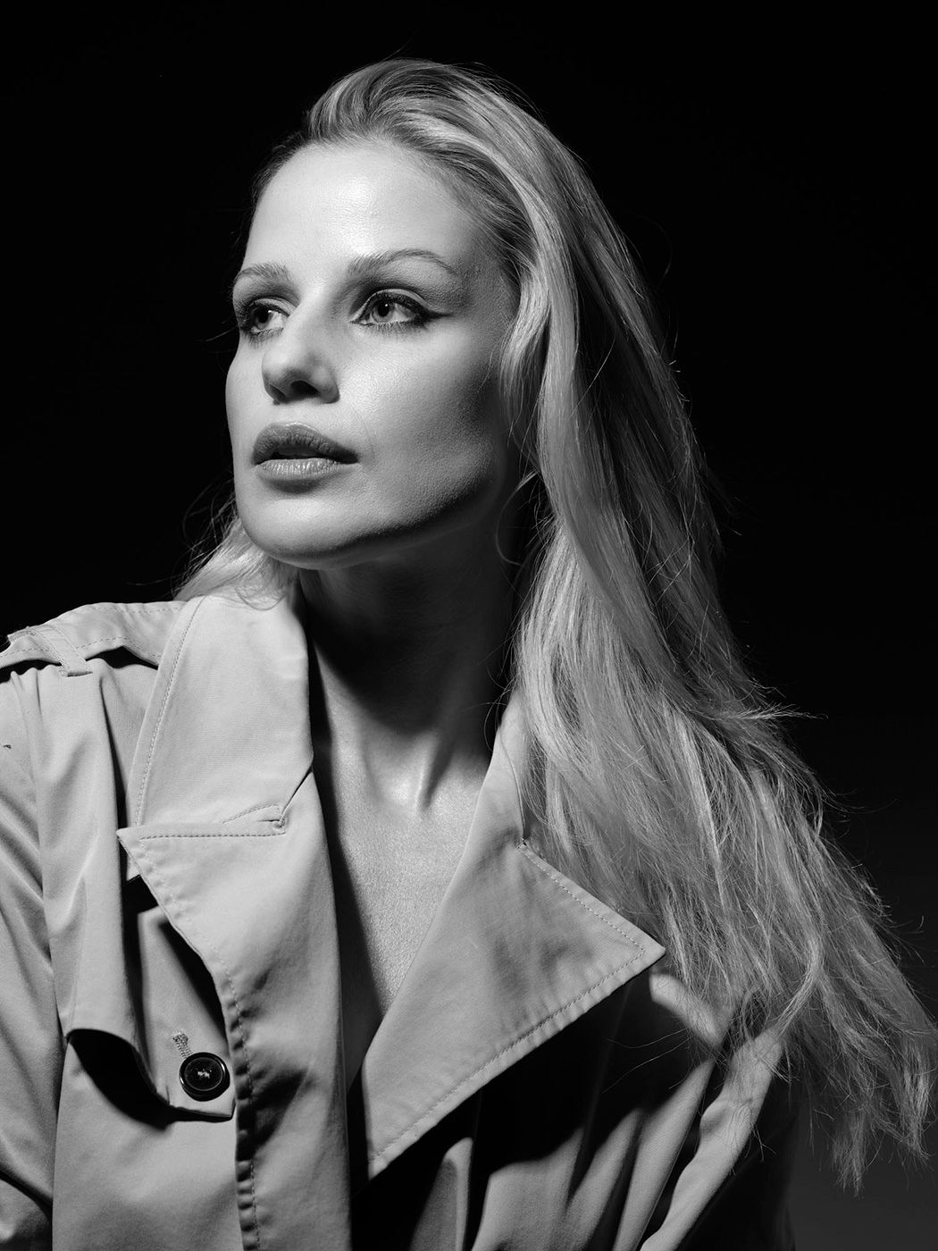 Black and white portrait of a blonde woman in a trench coat gazing upward against a dark background.