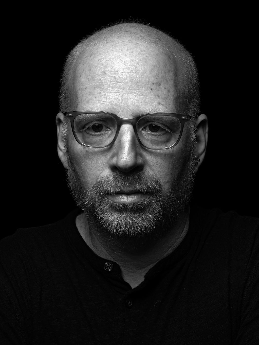 Black and white portrait of a bald man with glasses and a beard wearing a dark shirt. Oz Noy