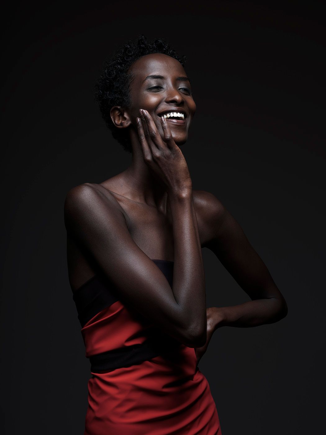 A joyful Black woman in a red strapless dress laughs elegantly against a dark studio background.