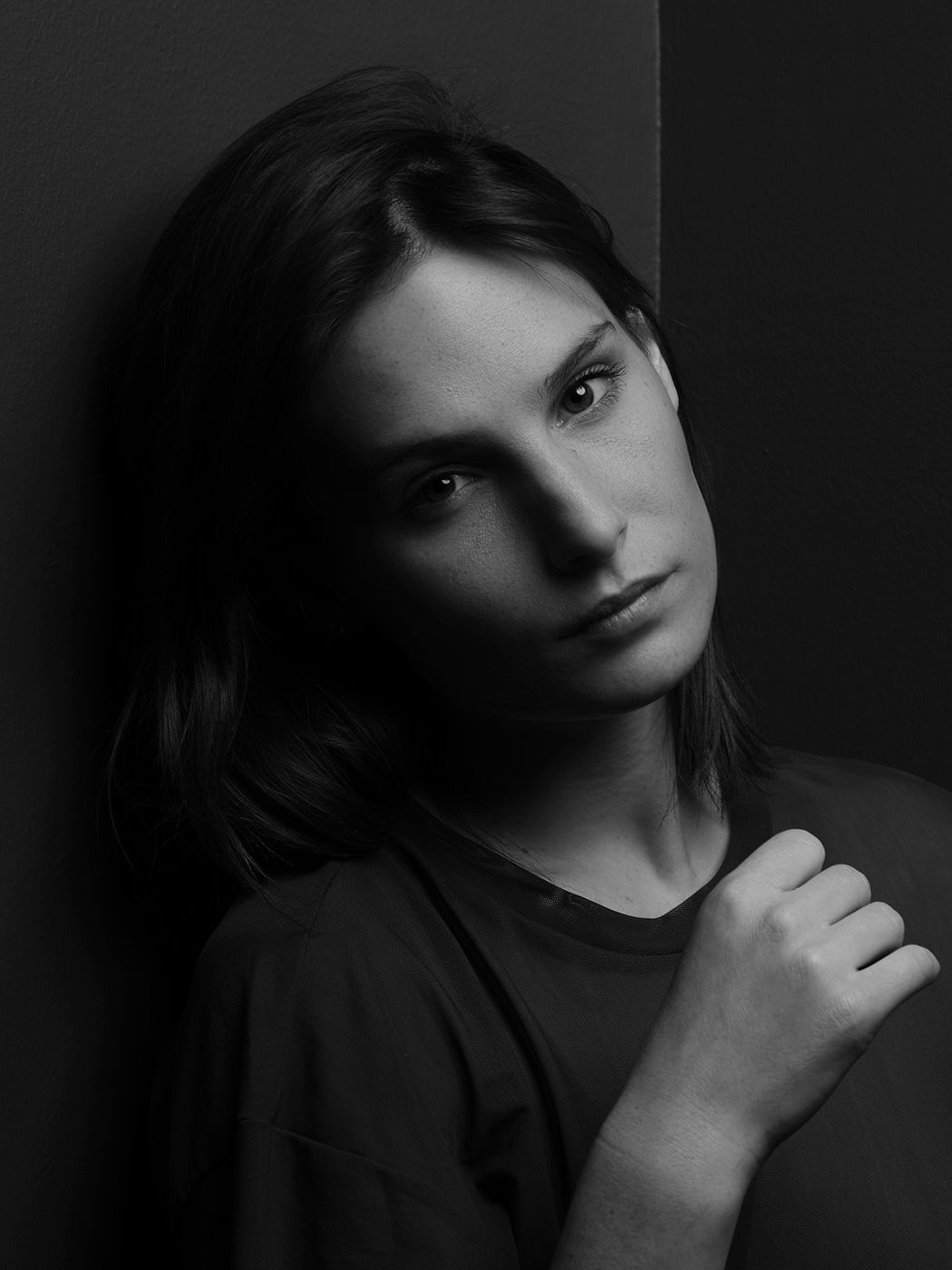 Black and white portrait of a young woman leaning against a wall, hand raised to chest, moody dramatic lighting.