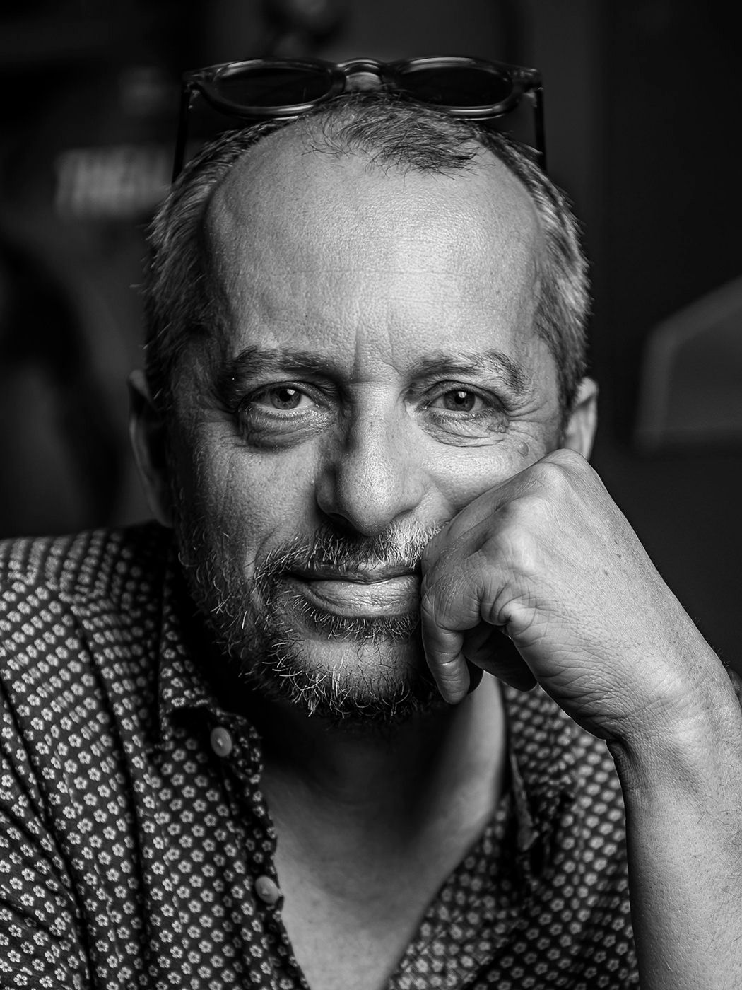 A smiling bearded man rests his chin on his hand in a candid black and white portrait photo.