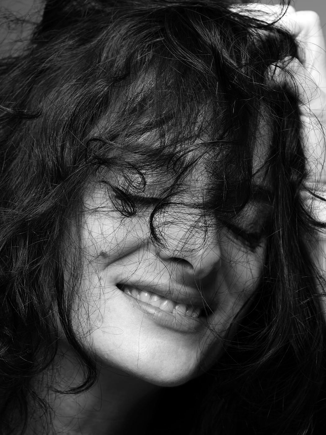 Black and white close-up of a smiling woman with dark curly hair blowing across her face.