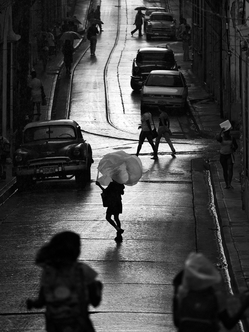 Black and white photo of pedestrians crossing a rain-slicked cobblestone street with vintage cars parked nearby.