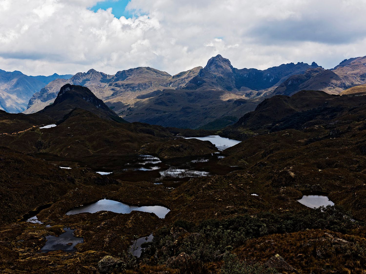 Panoramic view of Andean mountain peaks with scattered alpine lakes and wetlands under a cloudy sky.