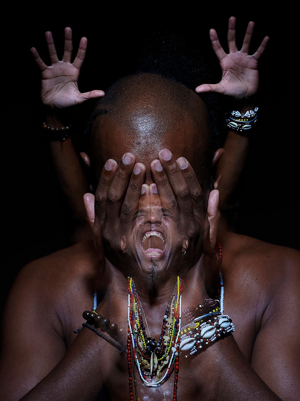 Shirtless man wearing colorful beaded necklaces covers his eyes while screaming, with hands raised behind his head. Omar Sosa