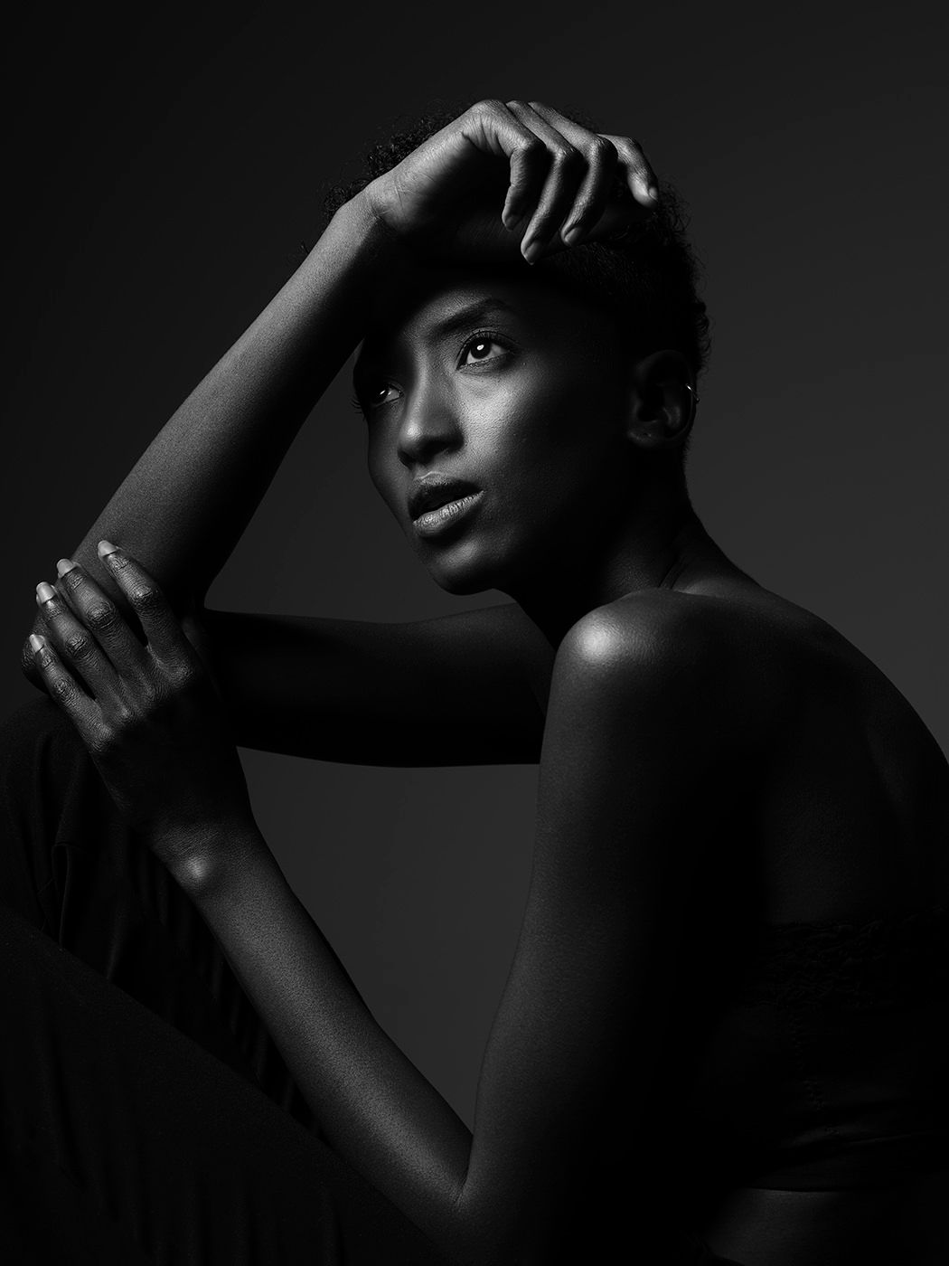 Black and white studio portrait of a young woman posing gracefully with hands near her face against dark background.