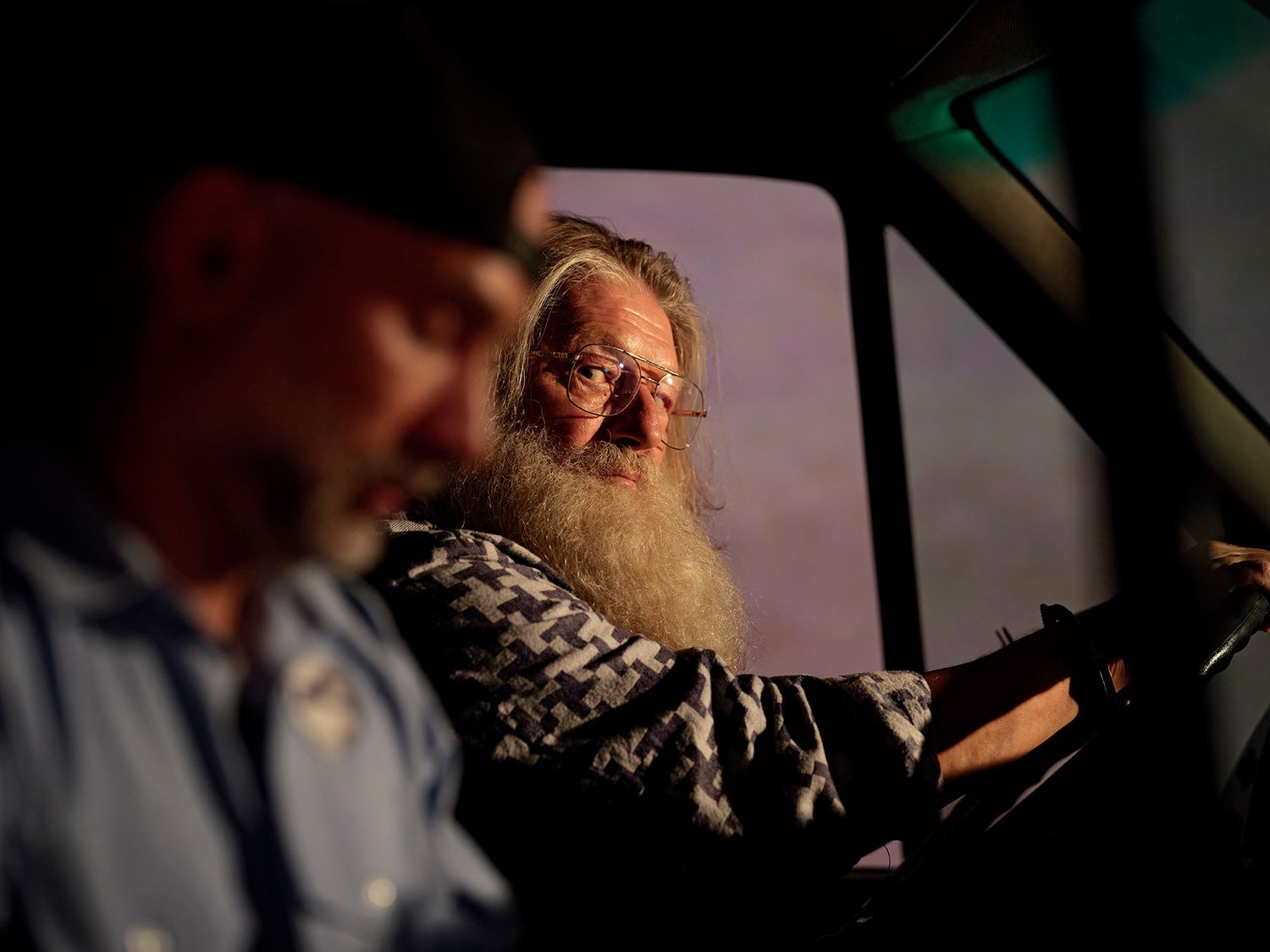An elderly bearded man drives a vehicle while a passenger looks on in warm evening light.