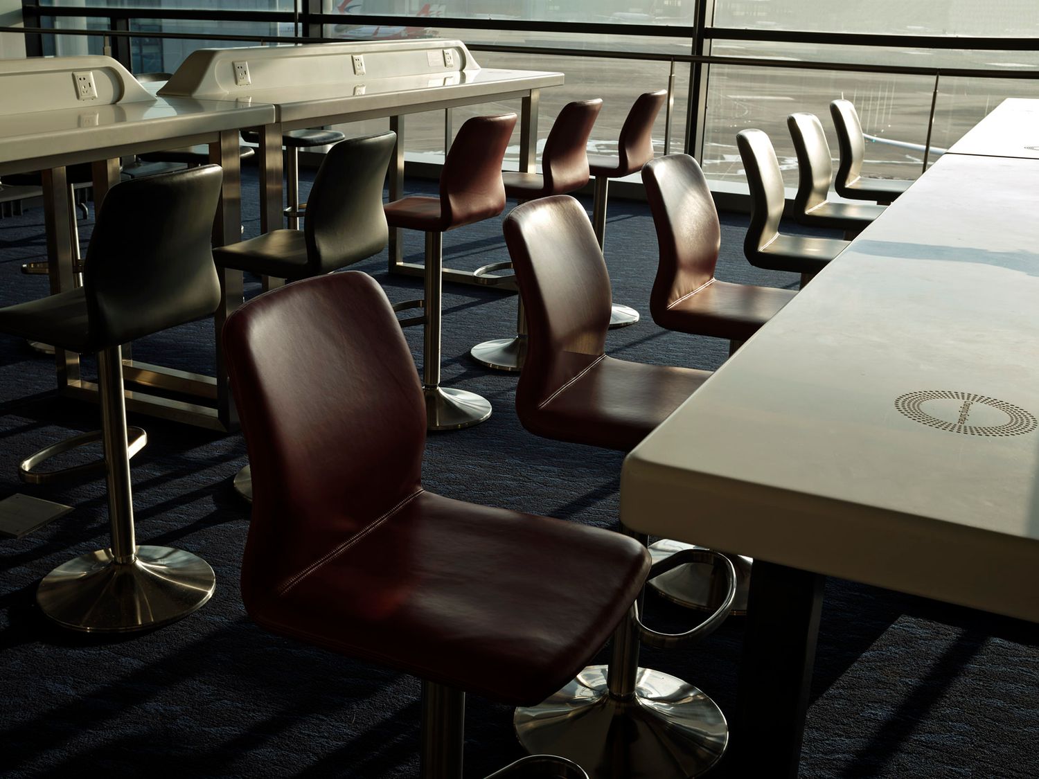 Modern airport lounge with dark leather swivel chairs and long white tables in sunlight.