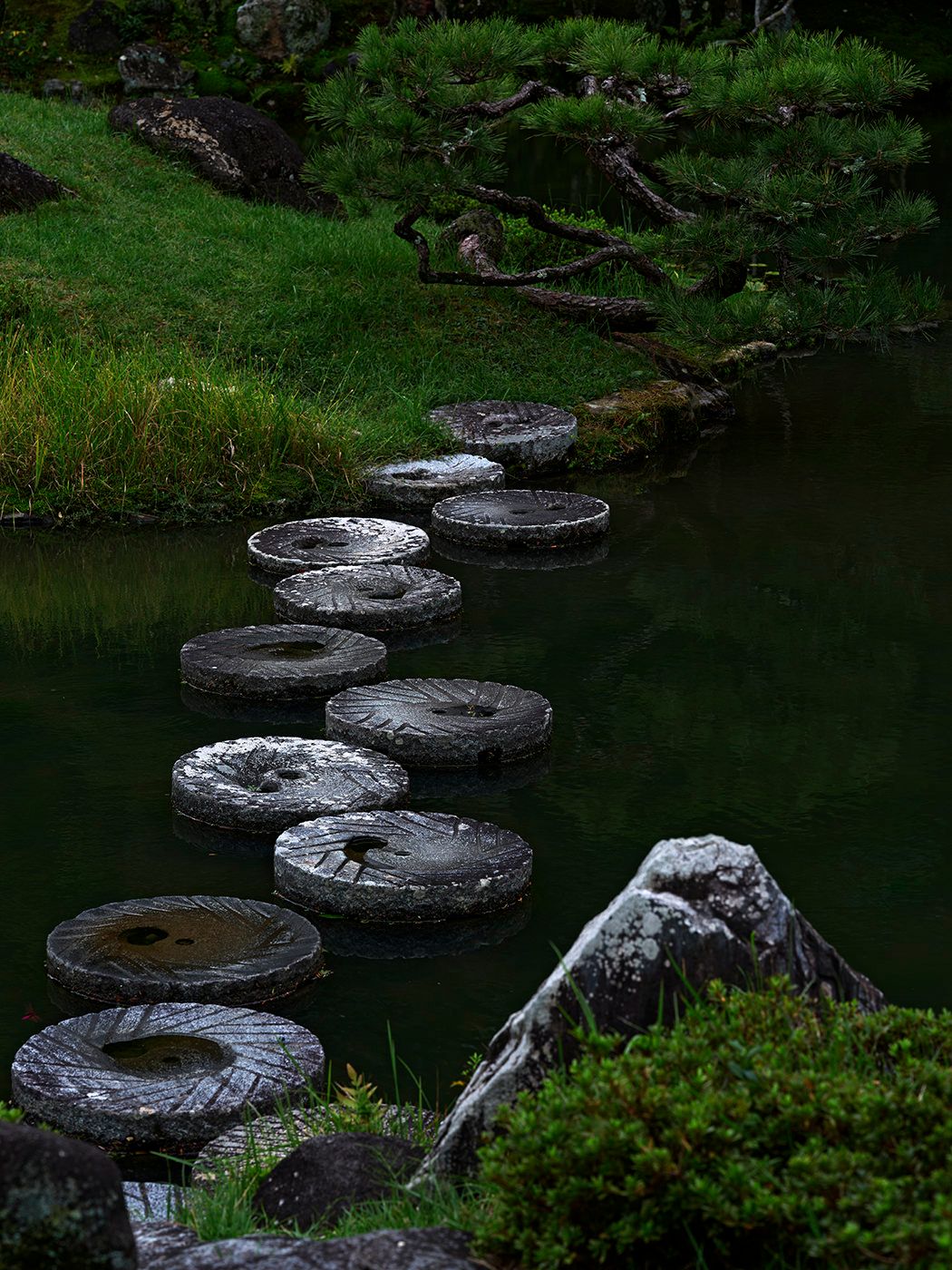 Circular stepping stones path across a serene dark green pond in a traditional Japanese garden.