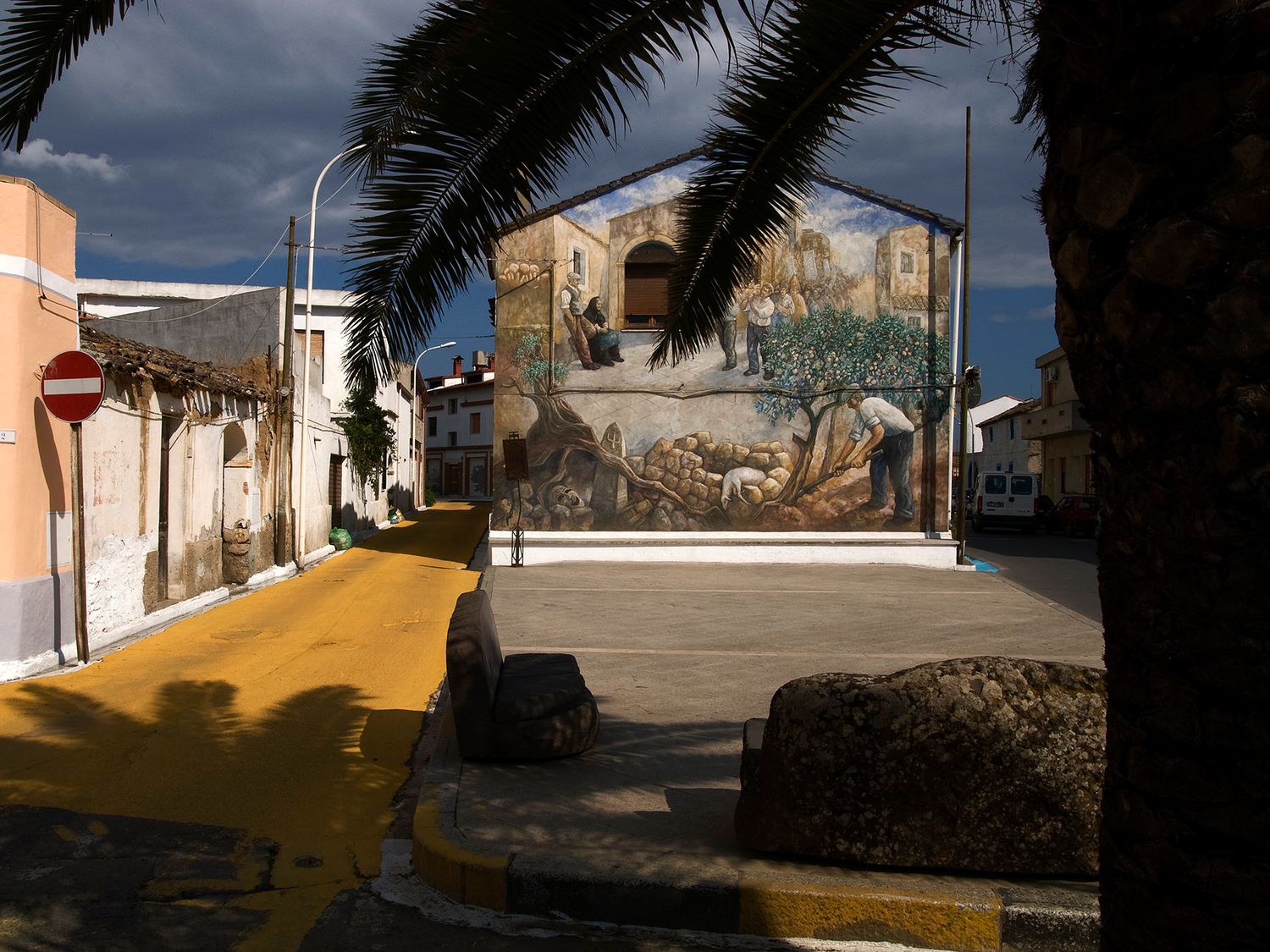A vibrant mural on a building facade in a quiet Mediterranean plaza, framed by palm trees and dramatic cloudy skies.
