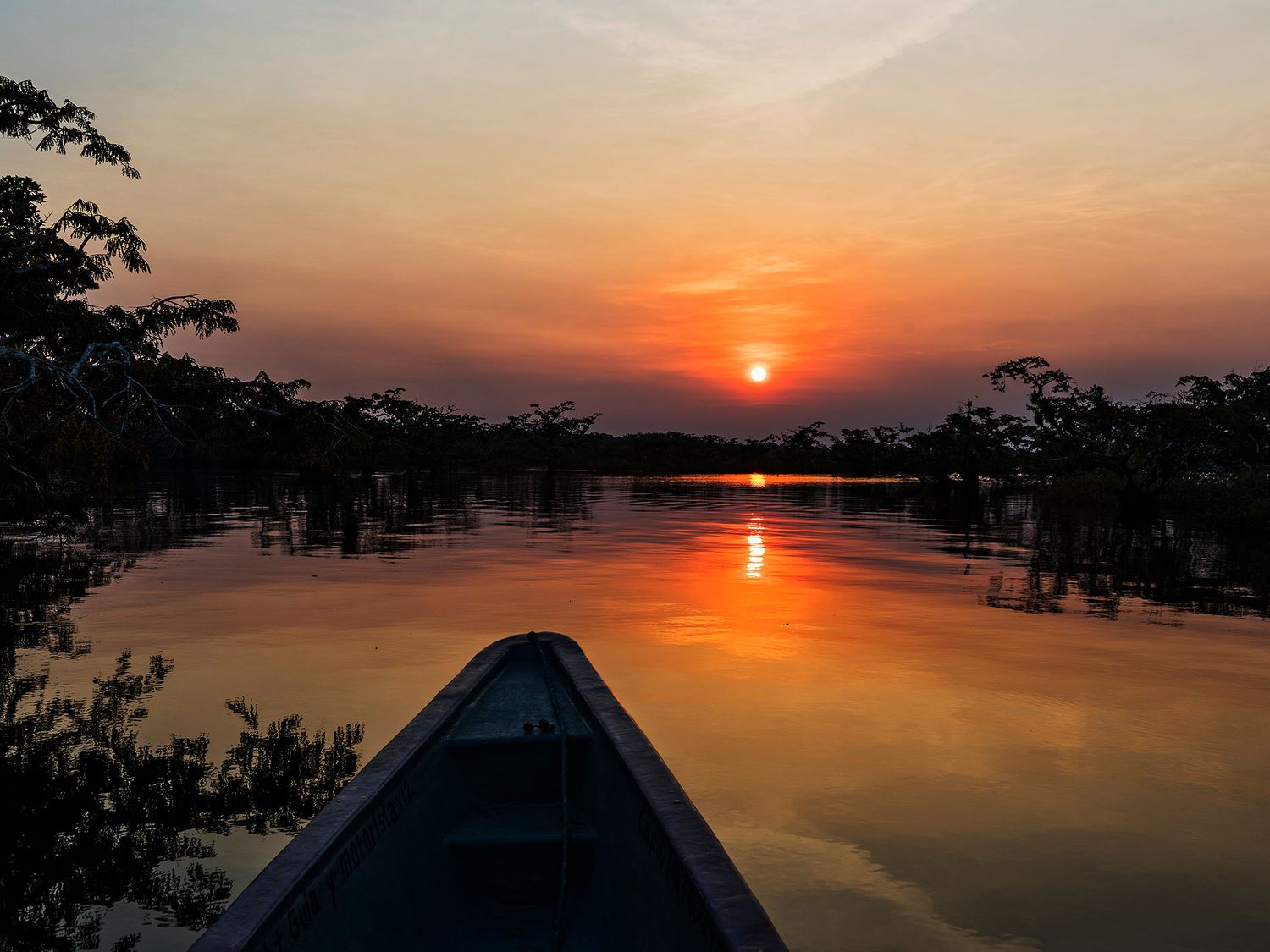Boat bow gliding on a calm river at sunset with golden reflections and silhouetted tropical trees.