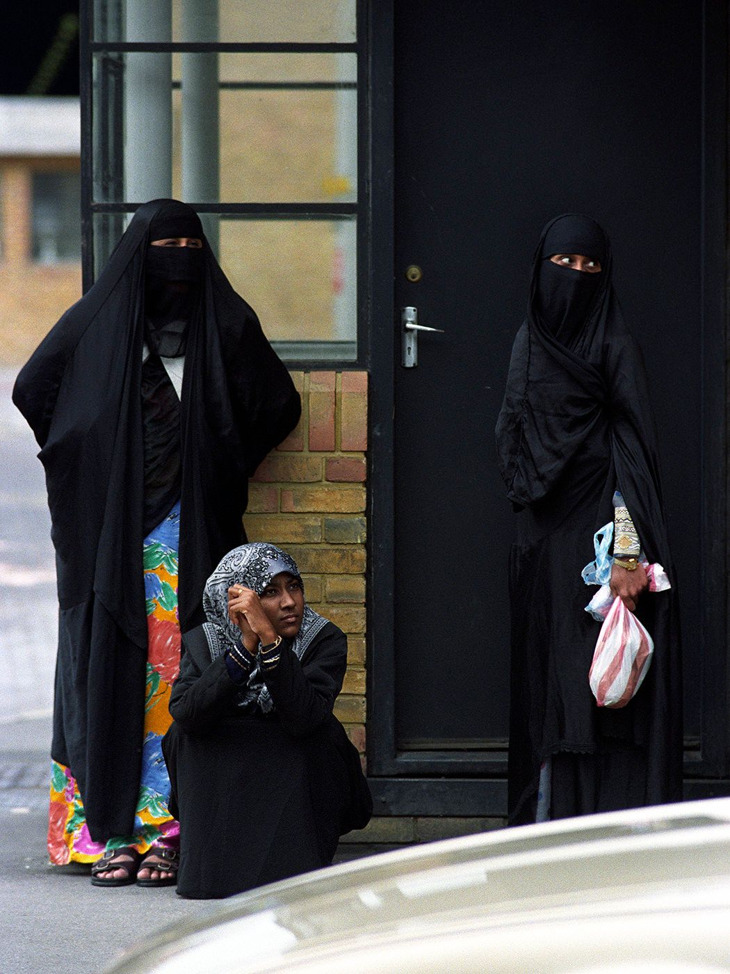 Two women in black niqabs and a child stand near a building entrance on a city street.