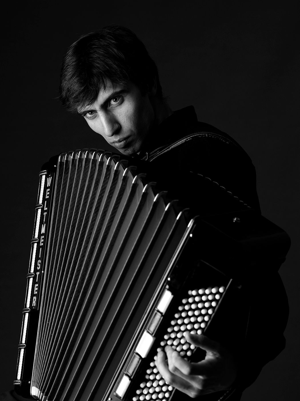 Black and white portrait of a young male musician playing a classic accordion against a dark background. David Yengibarian