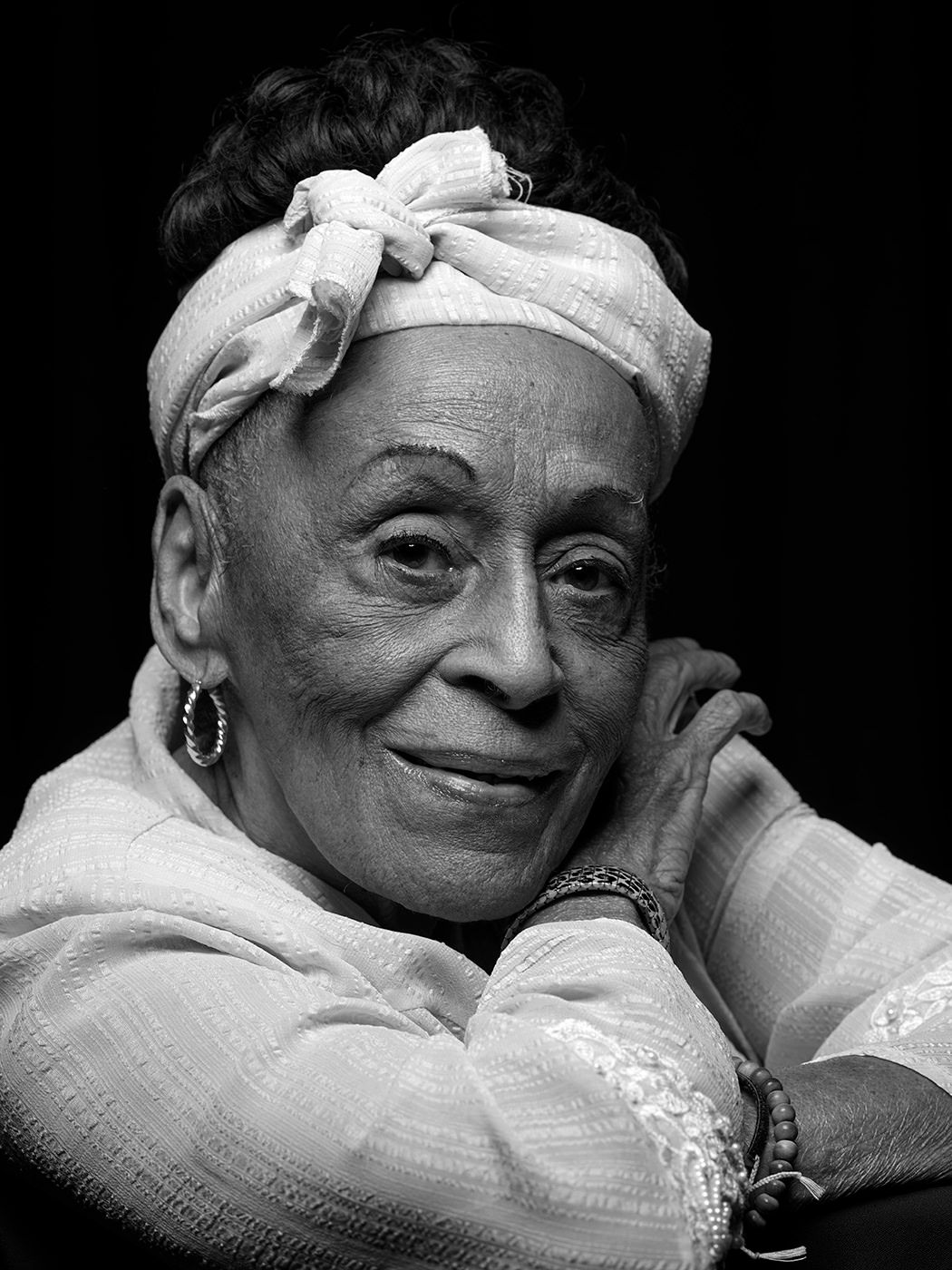 Elderly woman smiles warmly wearing a white headband and hoop earrings in a black and white portrait. Omara Portuondo