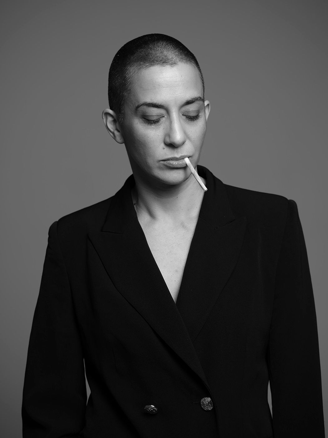 Black and white portrait of a person with shaved head wearing a black blazer with a cigarette in their mouth.