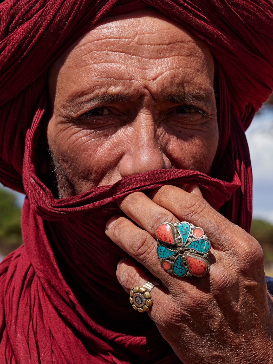 Close-up portrait of a man in red turban holding cloth to face wearing ornate rings.