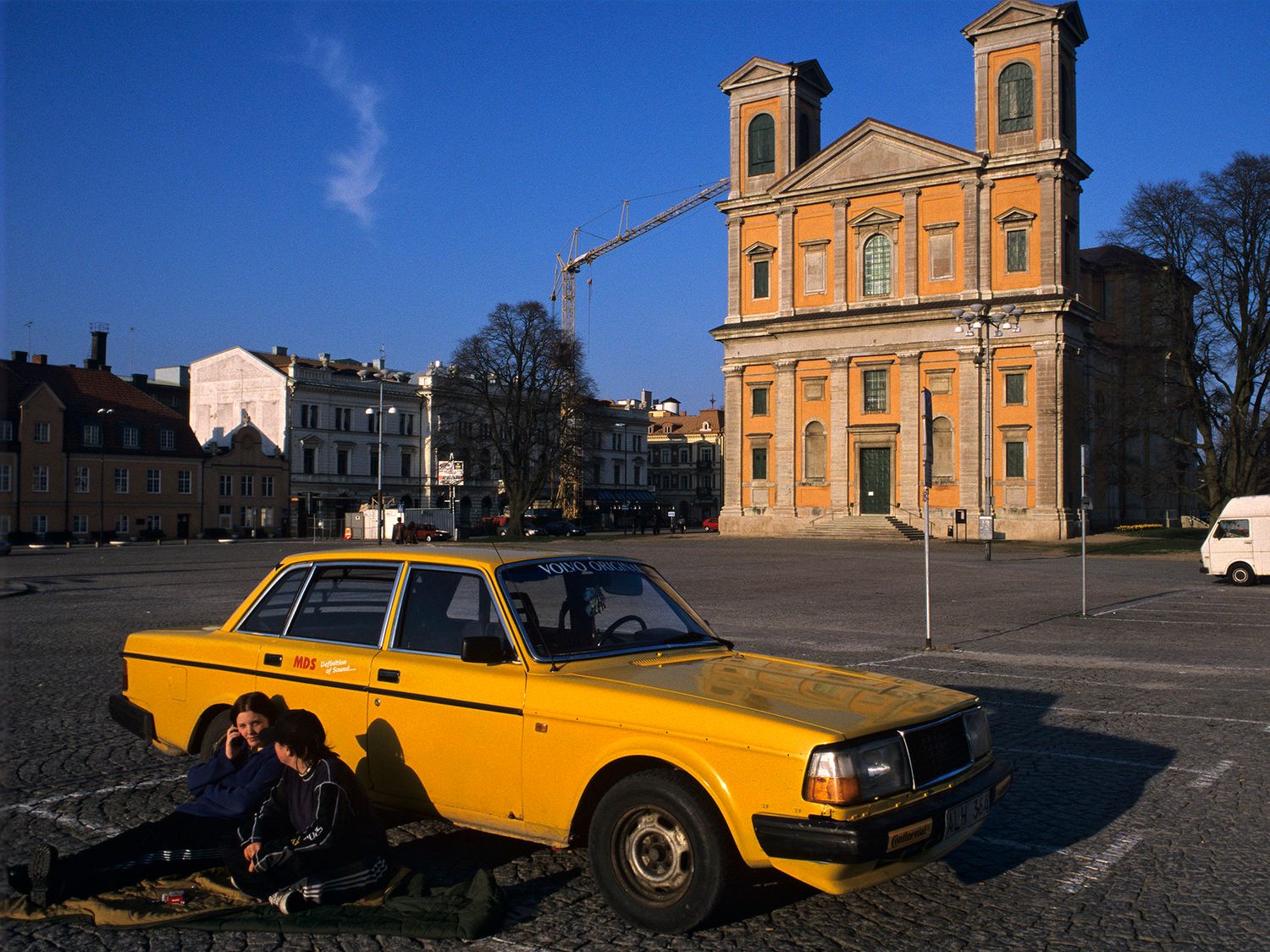 Yellow vintage Volvo car parked in a European town square with a grand baroque church in the background.