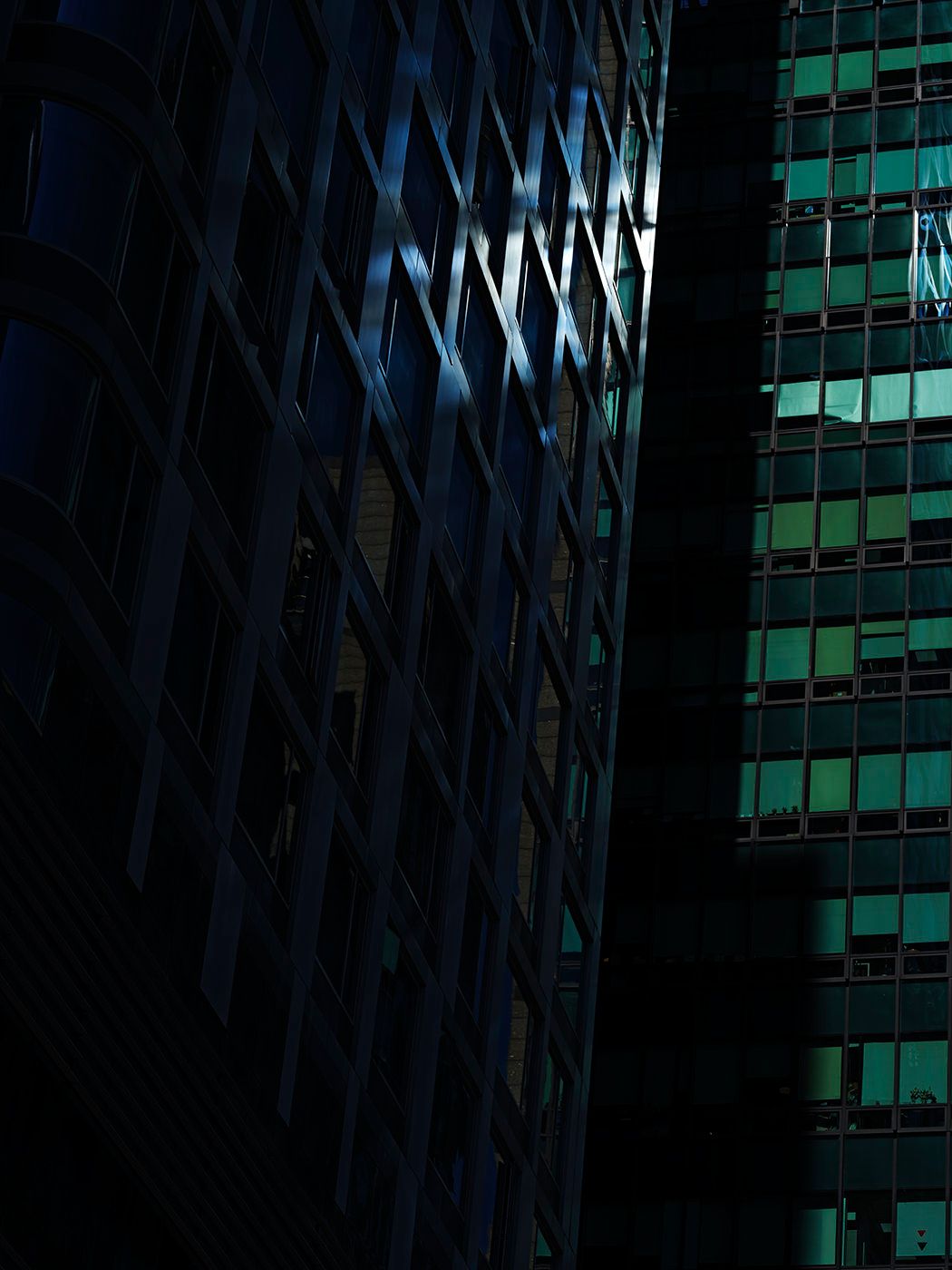 Dark urban skyscrapers at night with teal and green illuminated windows creating dramatic contrast.