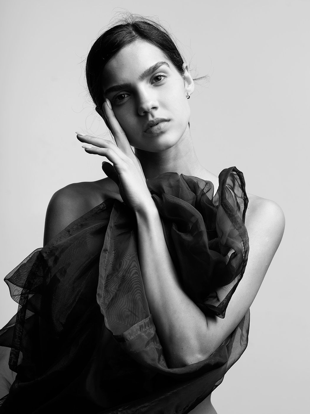 Black and white fashion portrait of a young woman in ruffled organza top, posing gracefully with hand near face.