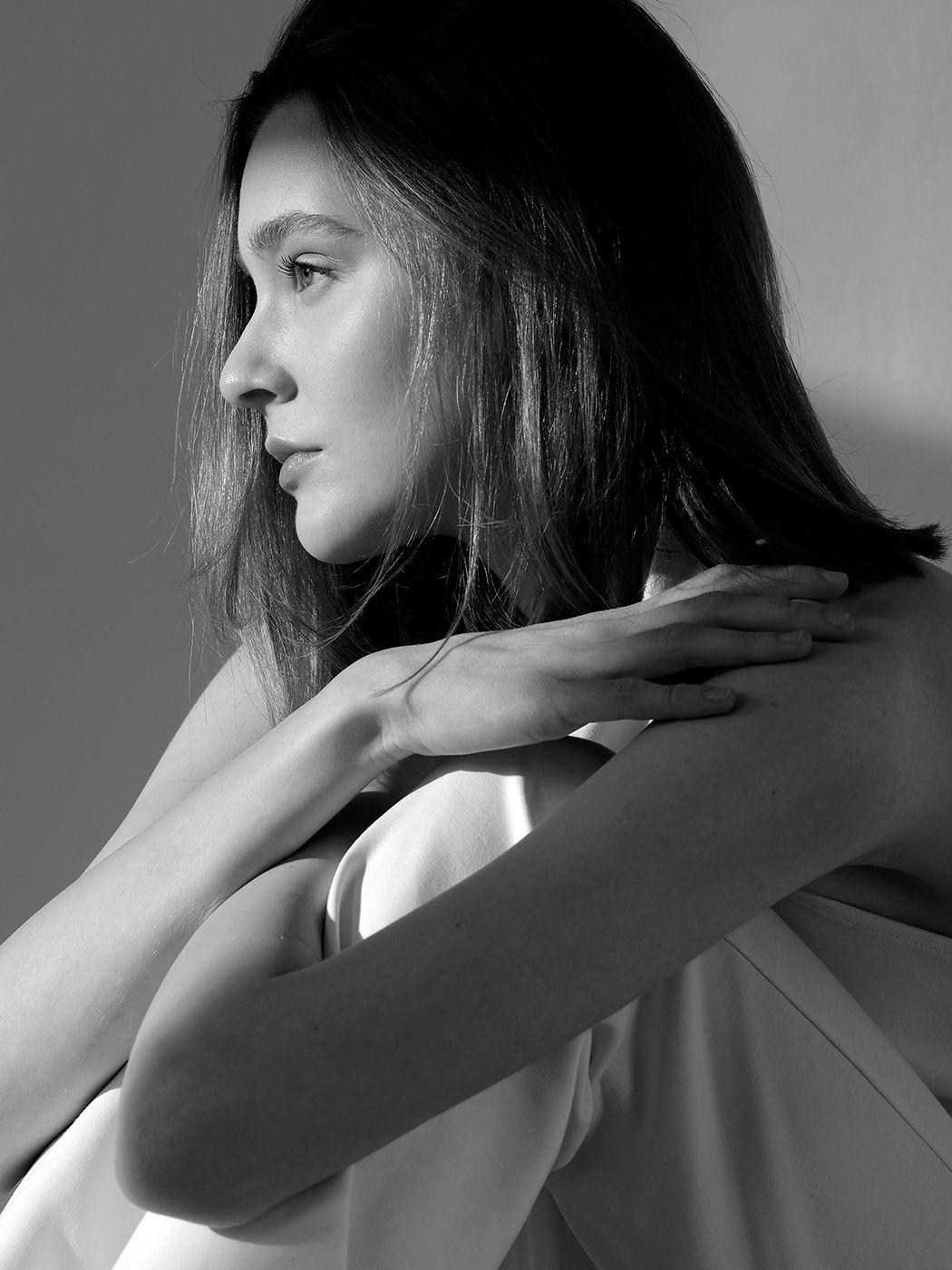 Black and white profile portrait of a young woman with long hair, arms crossed over her shoulder in soft studio lighting.