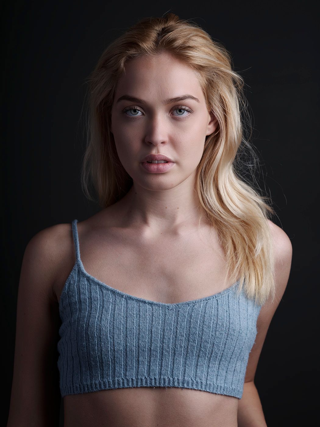 Young blonde woman in a light blue knit crop top poses against a dark background in a portrait photo.