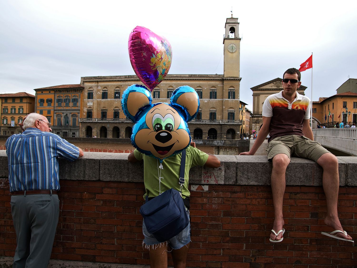 Mickey Mouse character with balloons poses on a bridge in an Italian piazza with historic buildings.