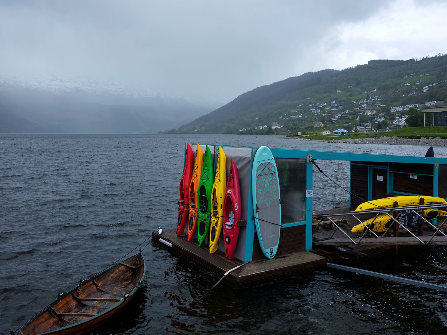 Colorful kayaks and paddleboards on a floating dock at a misty mountain lake on a cloudy day.