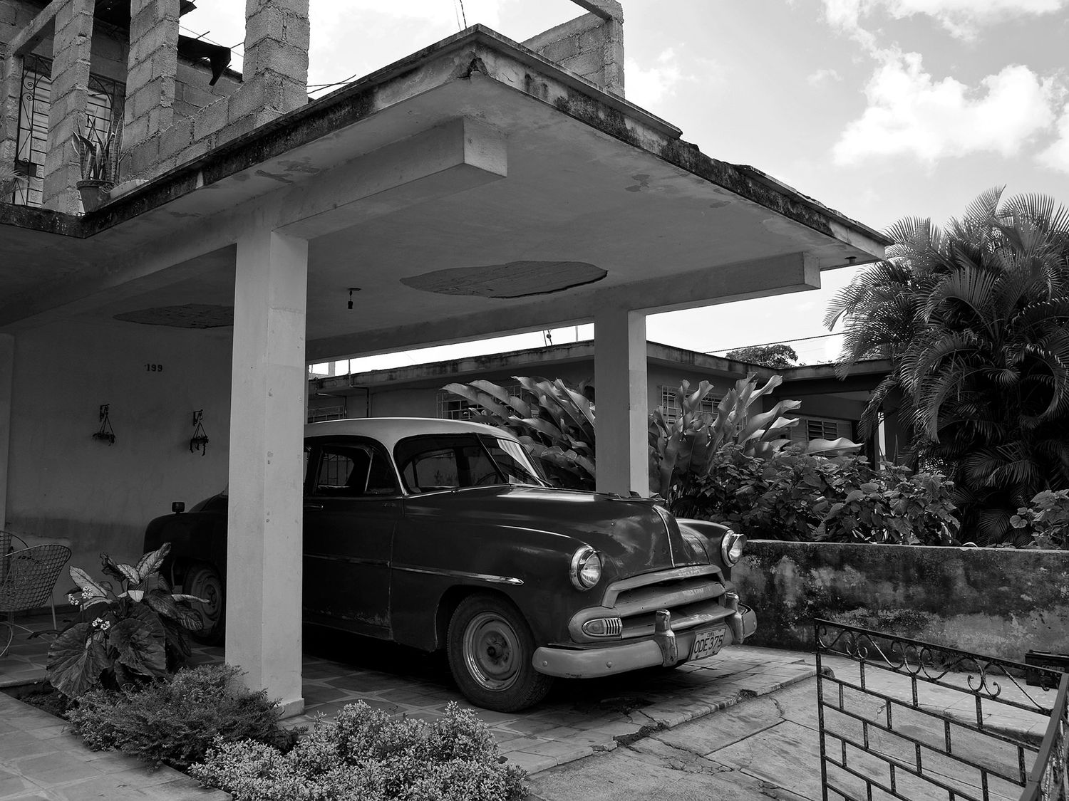 Vintage classic car parked under a covered carport beside a tropical home, black and white photography.