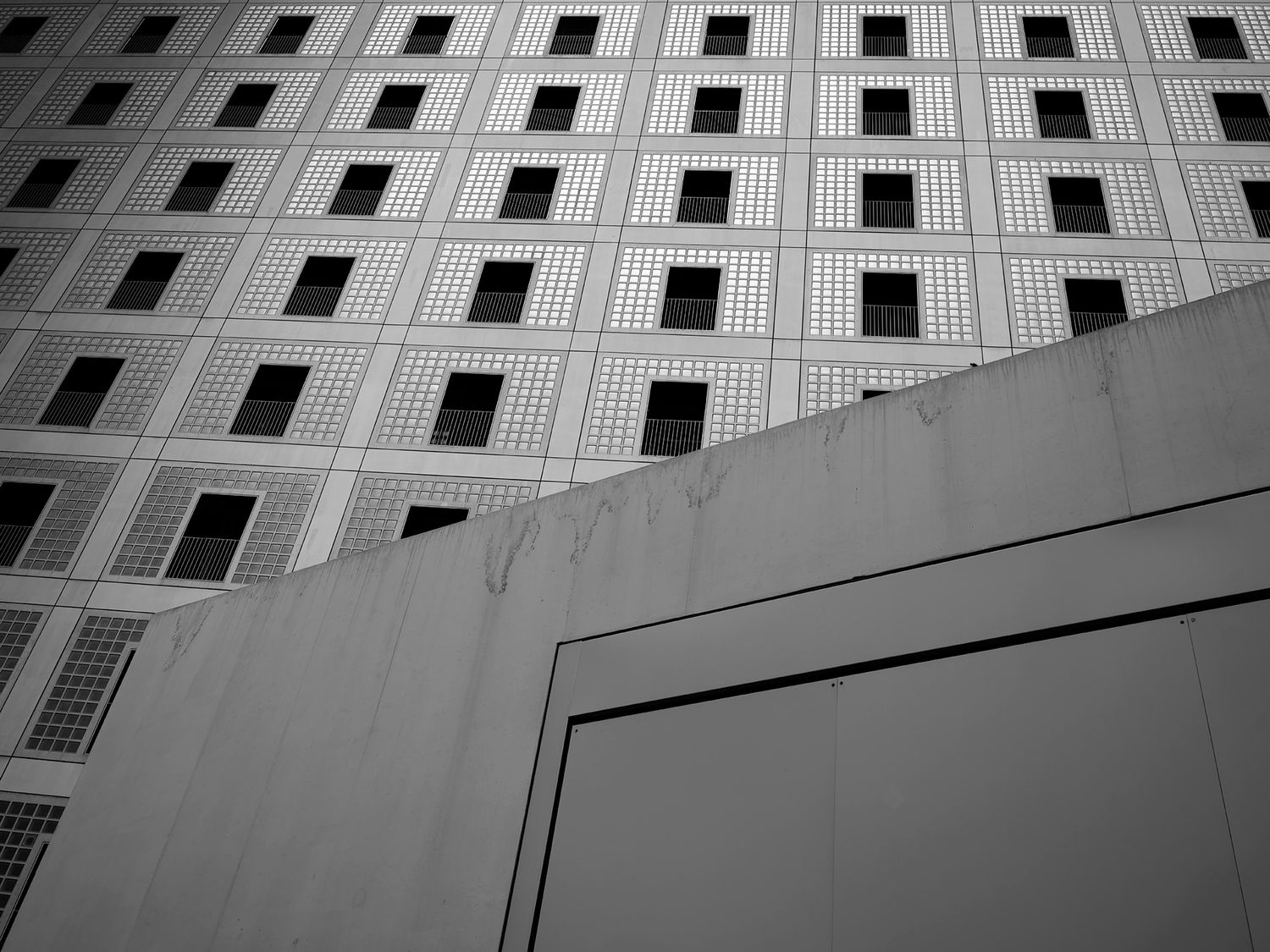 Black and white architectural photo of a tall building facade with grid windows beside a concrete wall.