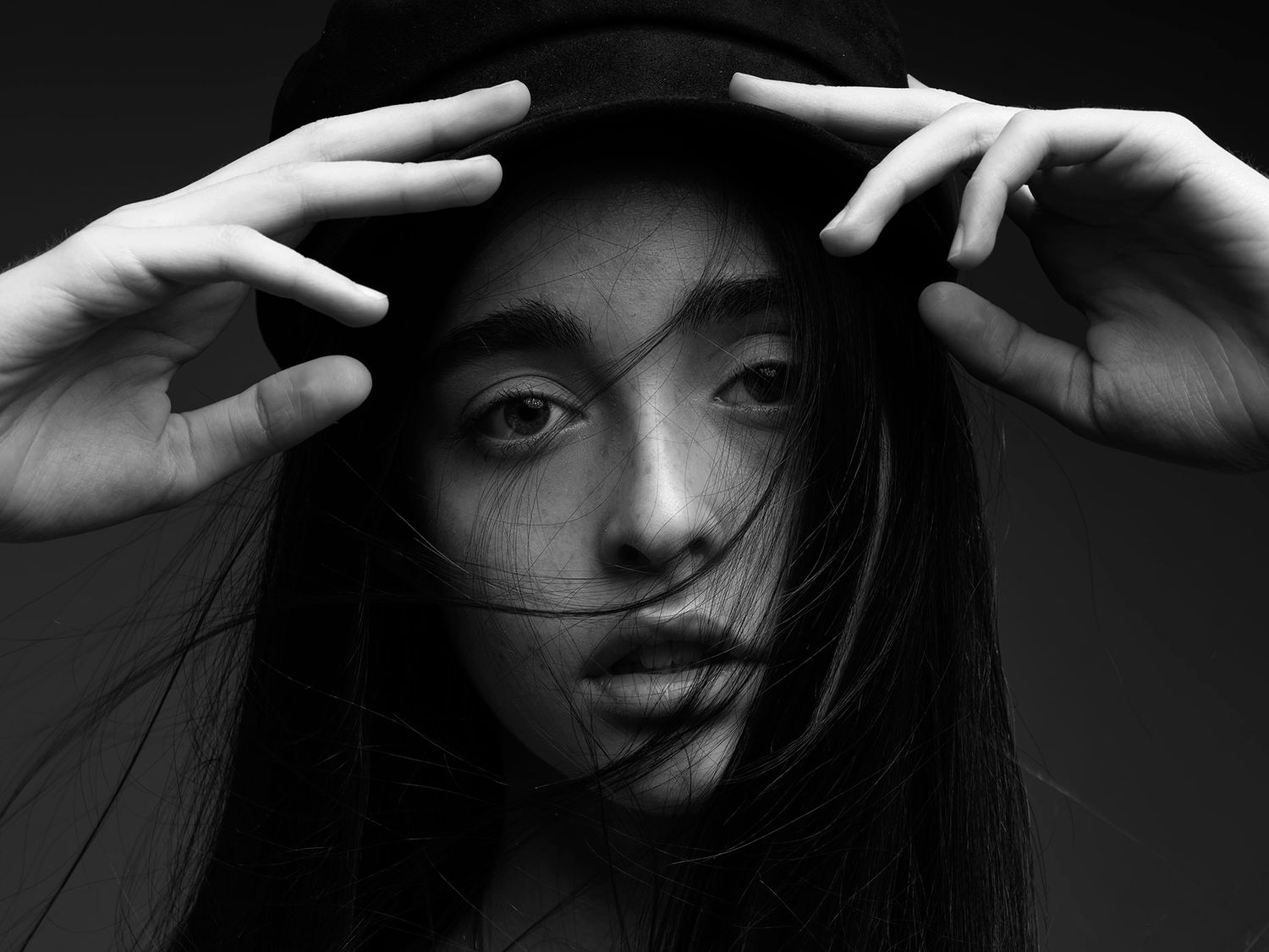 Black and white portrait of a young woman holding a dark cap over her head with wind-blown hair covering her face.