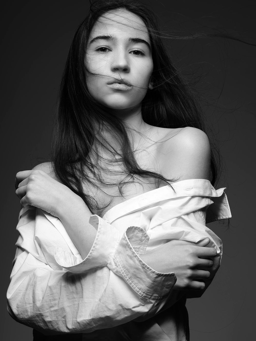 Black and white portrait of a young woman with windswept hair wearing an off-shoulder white shirt.
