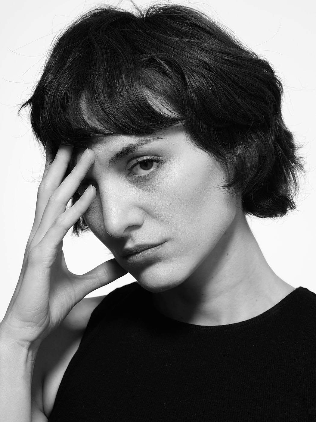 Black and white portrait of a woman with short dark wavy hair wearing a black top, hand raised to her face.