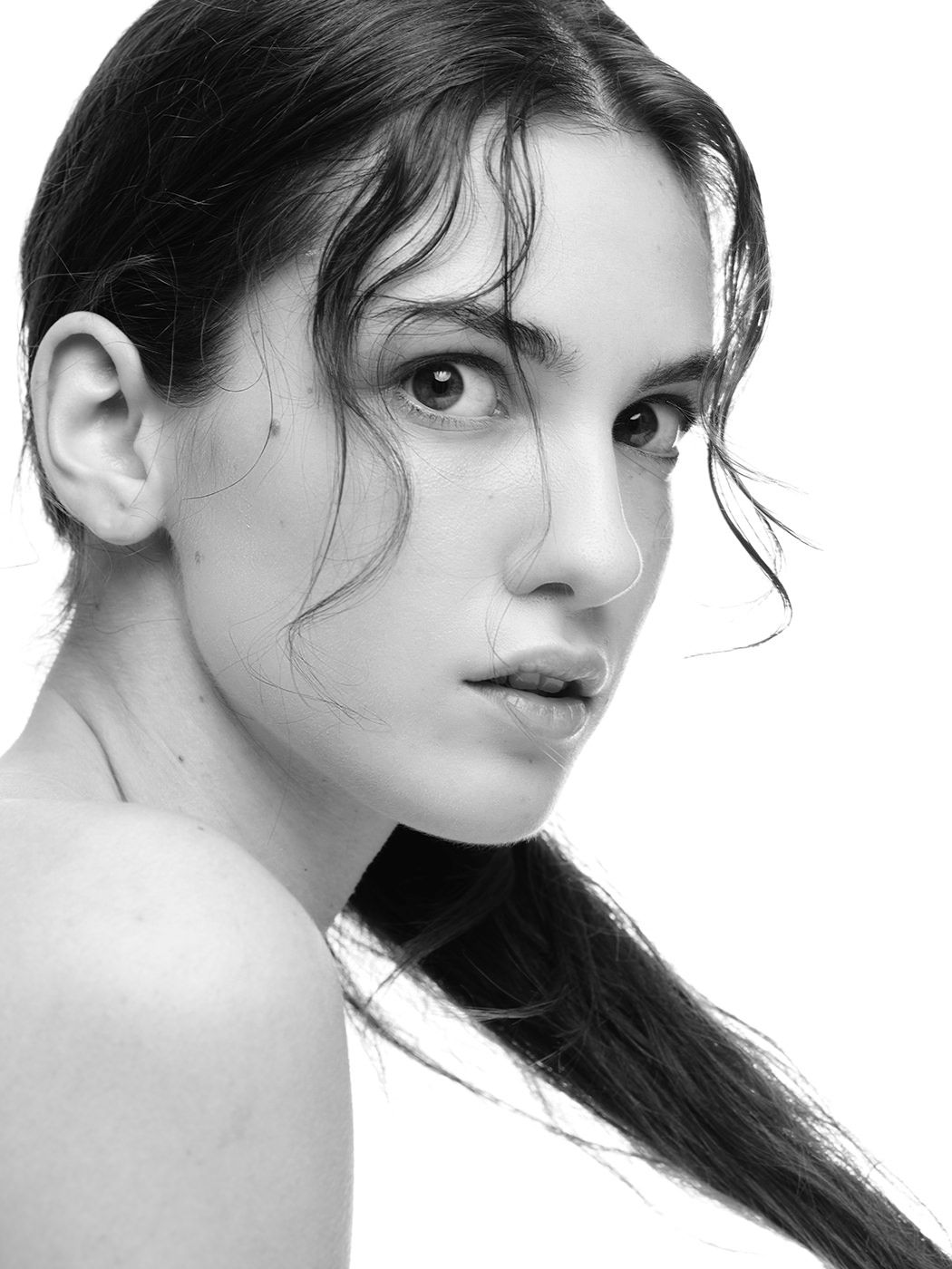 Black and white portrait of a young woman with wet hair, bare shoulder, and natural makeup, gazing over her shoulder.