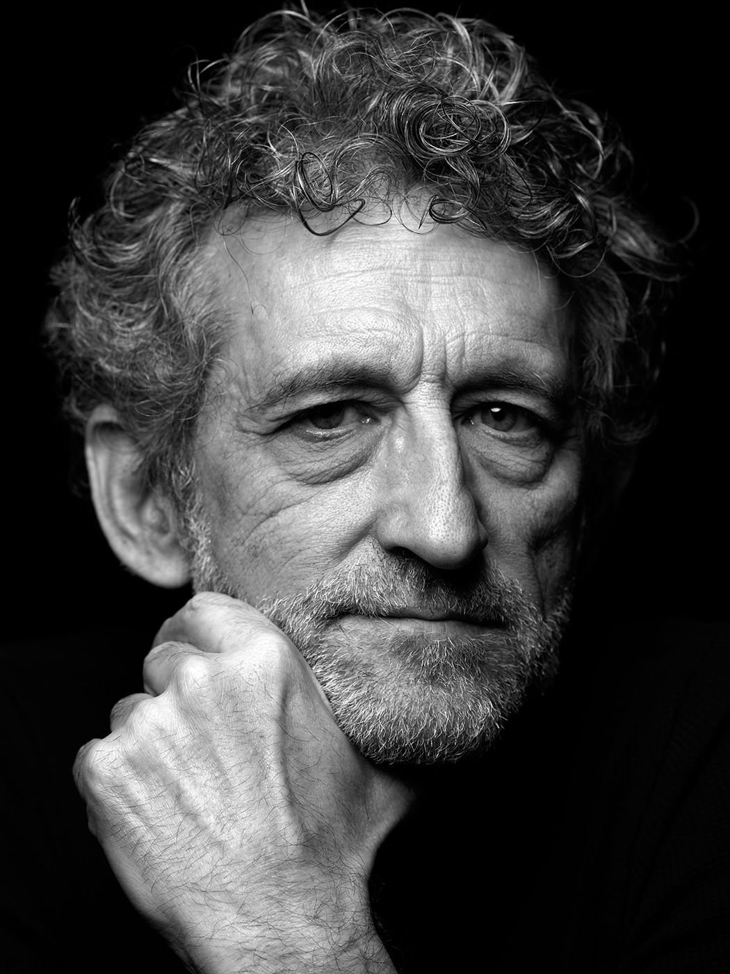 Black and white portrait of an older man with curly gray hair resting his chin on his fist against a dark background. Jojo Mayer