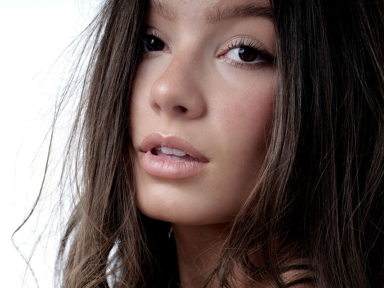 Close-up portrait of a young woman with long brown hair and natural makeup against a white background.