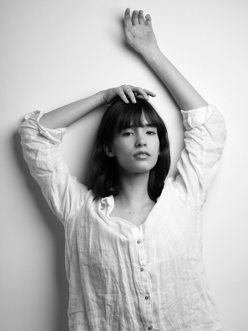 Black and white portrait of a young woman in a white linen shirt, arms raised gracefully above her head.