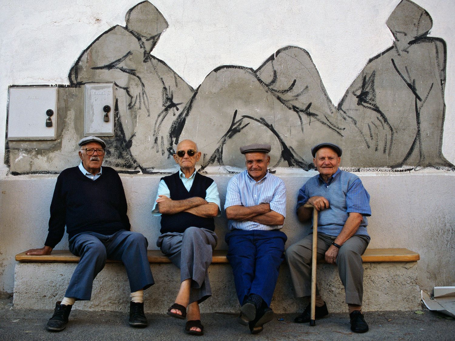 Four elderly men sitting on a bench in front of a mural depicting seated figures on a white wall.