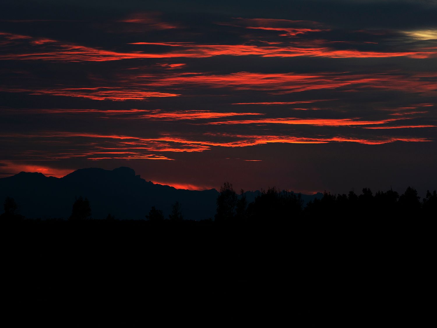 Dramatic red and orange sunset sky with silhouetted mountain and treeline against dark clouds.