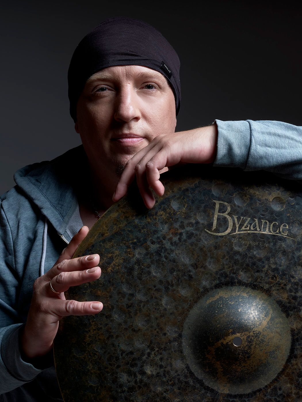 Musician poses with a Byzance cymbal wearing a black beanie and gray hoodie in a studio portrait. Mogyoró Kornél