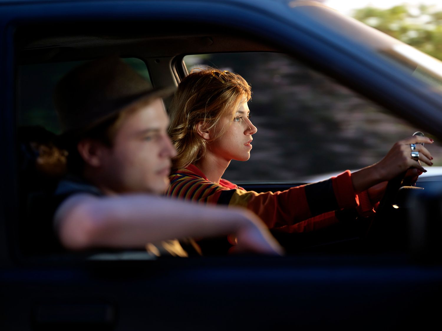 Color photo of two people inside a vehicle, a woman driving and a young man in the passenger seat.