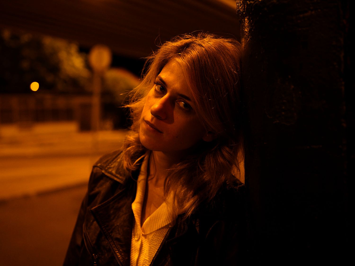 Young blonde woman leaning against a wall outdoors under warm orange night lighting.