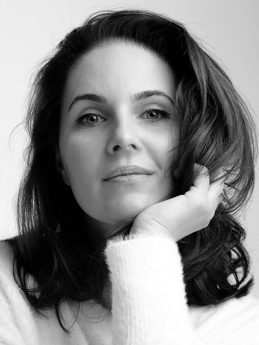Black and white portrait of a woman with shoulder-length dark hair wearing a white sweater, hand resting on her cheek.