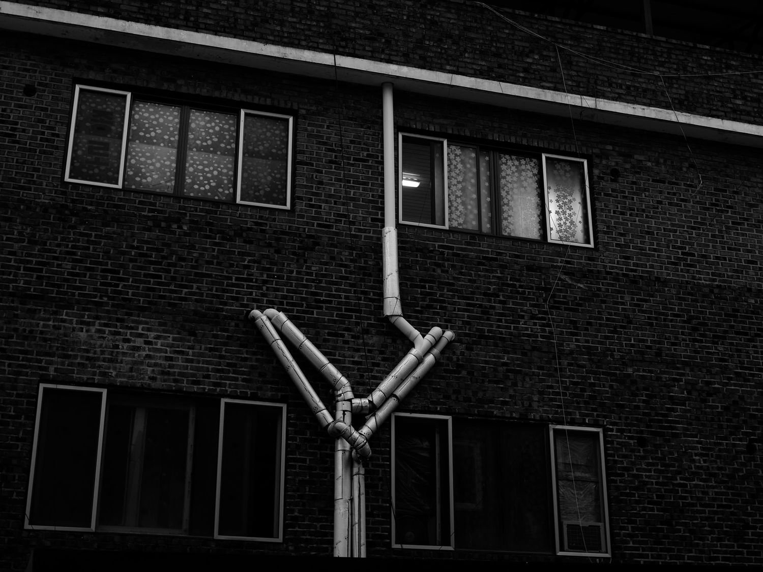 Black and white photo of industrial pipes forming a Y-shape on a dark brick building facade at night.