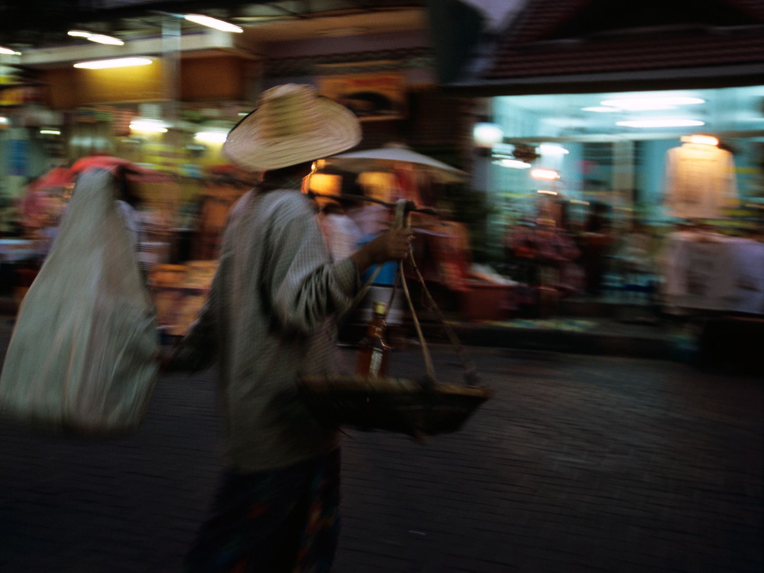 A street vendor wearing a traditional conical hat carries balance pole baskets through a busy night market.