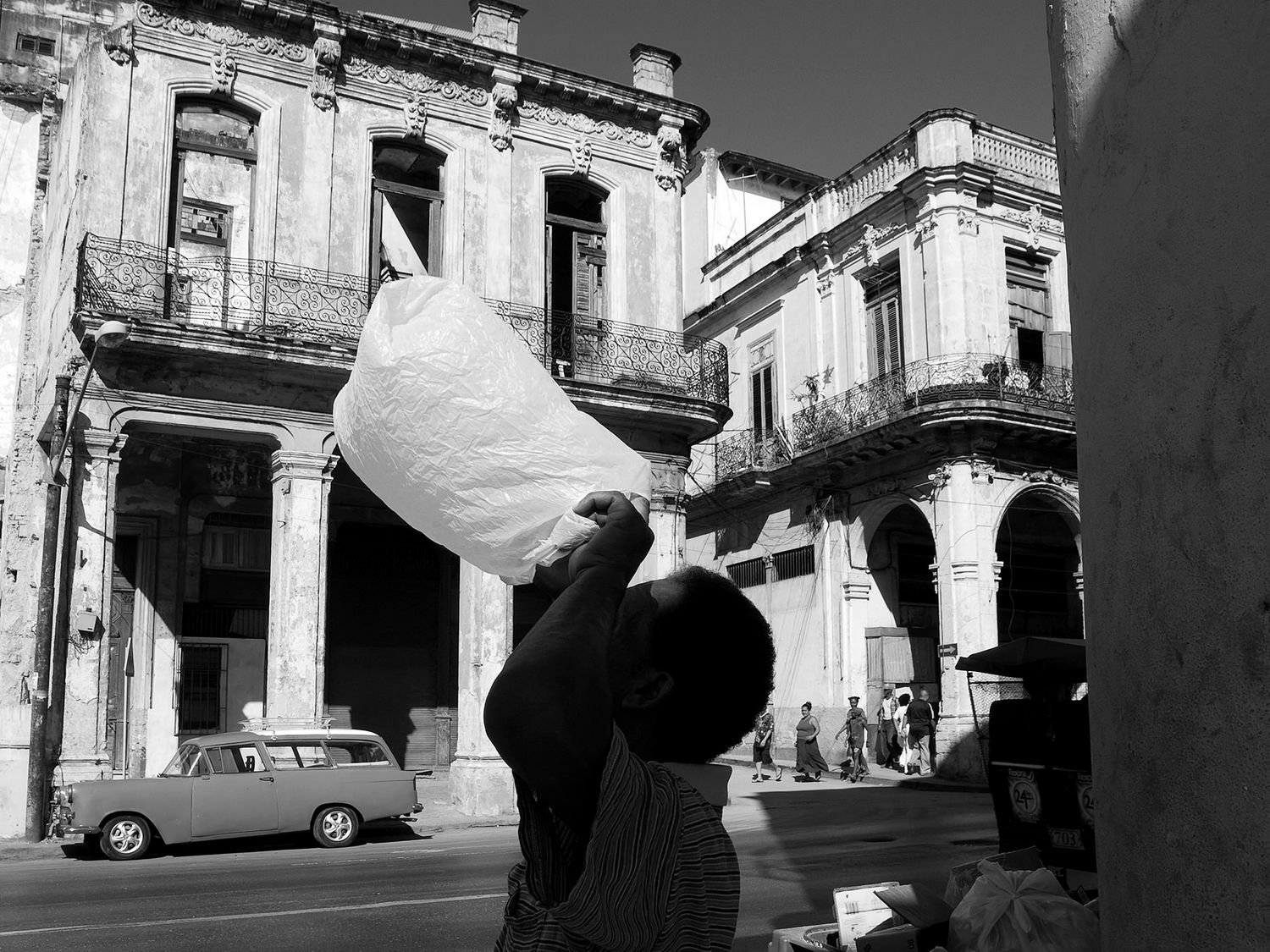 Black and white street scene in Havana, Cuba, with a vendor carrying a large bag near vintage cars and crumbling colonial buildings.