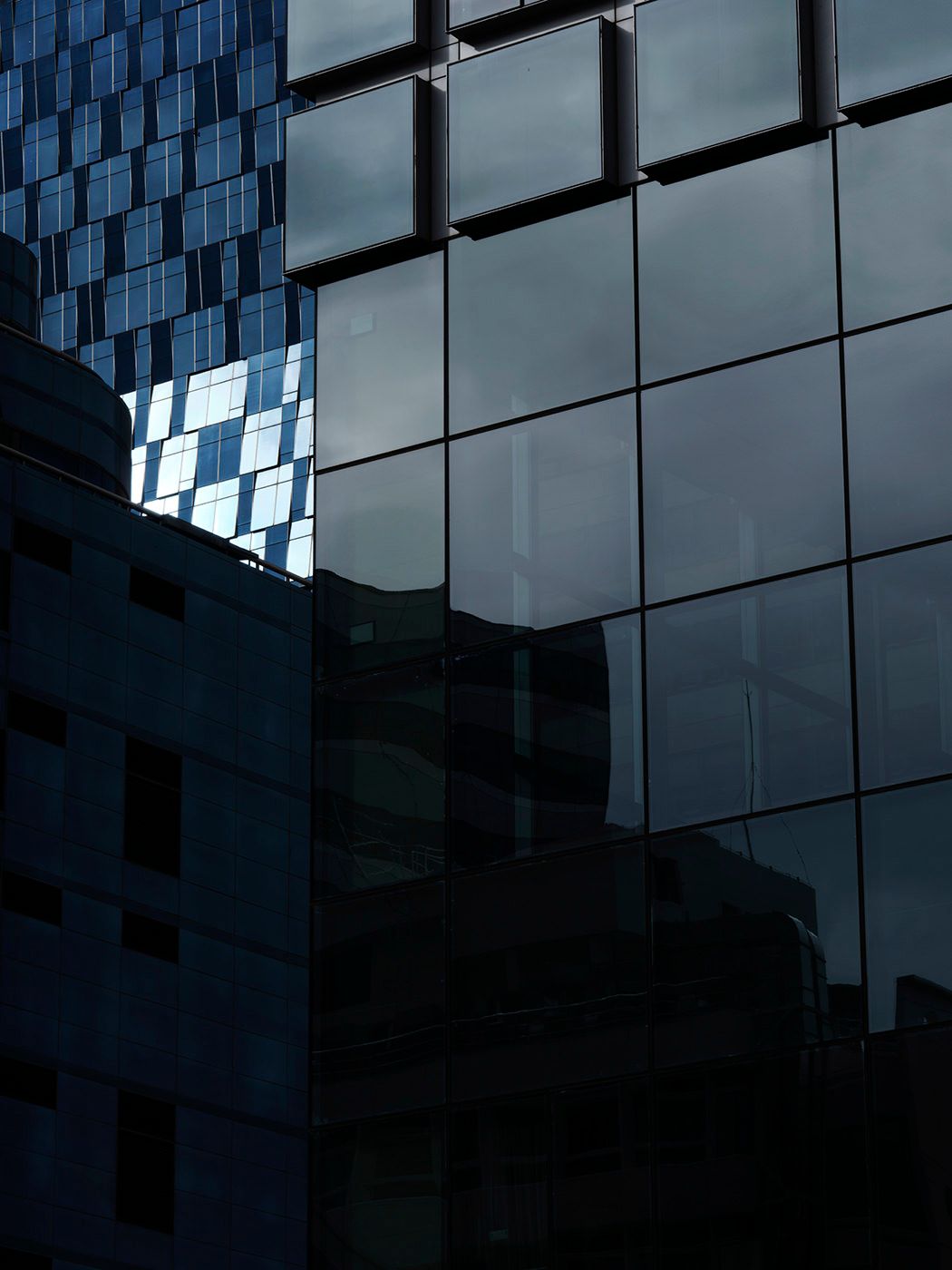 Dark glass facade of a modern skyscraper reflecting cloudy sky, with geometric grid patterns and blue checkered building nearby.